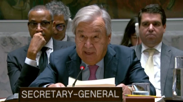 Guterres headshot of him speaking with a name plaque in front of him with the words secretary general on it. Two men sit behind him