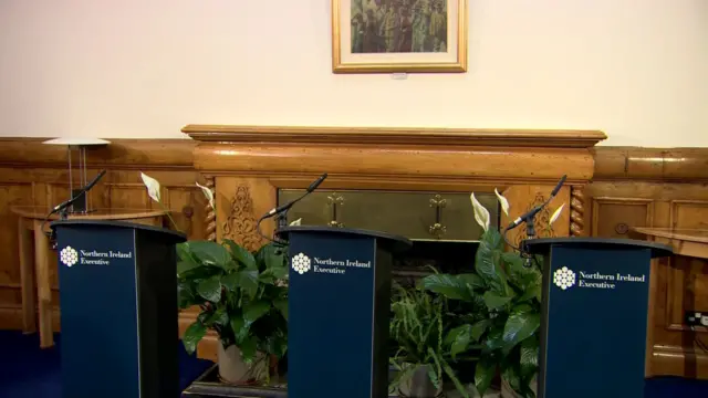 Three podiums with Northern Ireland Executive written on them in a room with a foreplace and wood panelling