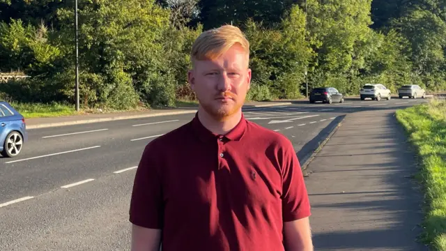 Daniel McCrossan stands on a footpath beside cars travelling on the A5 road. He is wearing a red, buttoned up polo shirt and is looking at the camera.