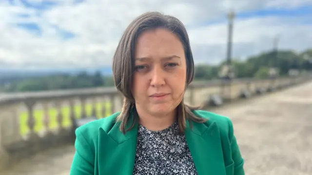 Deborah Erskine - a woman with shoulder-length brown hair looks at the camera. She is wearing a green suit jacket and a floral patterned shirt. She is stood on a stone-paved road outside of Stormont, with the background blurred.