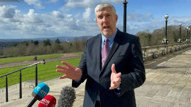 John O'Dowd is mid interview with three microphones in shit - a blue RTE branded one, a red BBC News NI branded one and a fluffy grey one. John has grey hair and a small fringe. He is wearing a blue shirt and pink tie under a navy suit jacket. He is talking with his hands in motion and is looking at the camera.