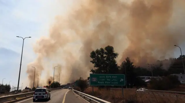 Large plumes of smoke over a highway with a road sign saying "Rosh Pina"