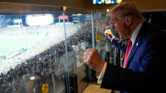 Donald Trump, wearing a suit, pumps his fists and shouts behind the glass window of his suite in a American football stadium