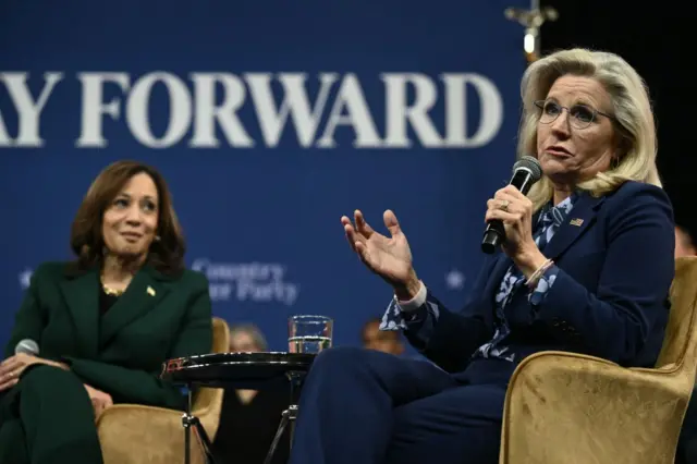 Kamala Harris and Liz Chenney sit on armchairs during a town hall event. Chenney is speaking into a mic, while Harris looks at Cheney. Harris is wearing a dark green suit, while Chenney is wearing a navy suit and floral blouse.