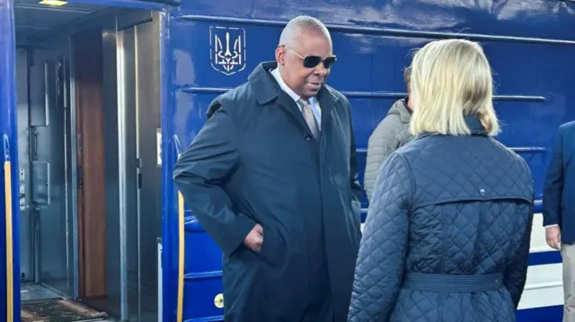 Lloyd Austin just stepped down of train as he arrives in Kyiv. He's wearing blue raincoat and blue trousers as well as sunglasses. He's speaking to US Ambassadoe Bridget Brink in blue raincoat. Blue train with Ukraine sigil in background