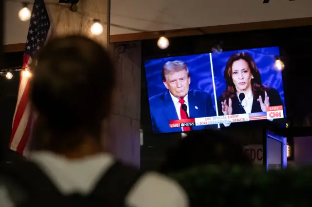 Blurred woman wearing white shirt and backpack in foreground watches TV broadcasting Trump/Harris debate