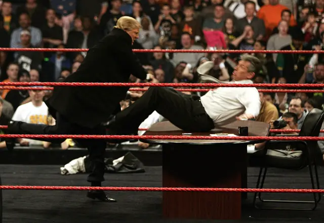 Donald Trump throws Vince McMahon across a table inside a WWE wrestling ring in 2007. Both men are dressed smartly in shirts and trousers, with Trump also wearing a blazer jacket. Crowds look on at the two.