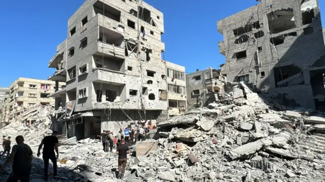 Residential block flattened to rubble surrounded by other damaged buildings, which people walking through it, in Beit Lahia