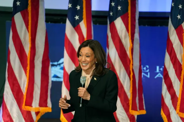 Kamala Harris campaigning. She's wearing a black suit with blush pink blouse with low ribbon around her neck and holding a mic in her left hand. Four American flag in blue background