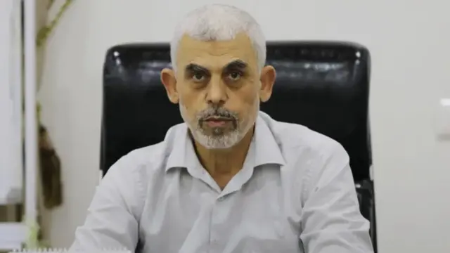 A headshot of Yahya Sinwar, wearing a white dress shirt, sitting in a black leather desk chair and staring into the camera