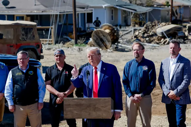 Former President Trump speaking at a press conference. He's wearing dark blue suit, white blouse and bright red tie. In background are six North Carolina officials observing him speak. Behind them are debris left by Hurricane Helene