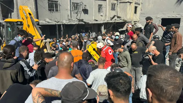 Huge crowd of people stand on rubble with emergency services worker holding a stretcher in the middle of them. There's a bright yellow digger amongst the crowd of people. Behind damaged building can be seen.