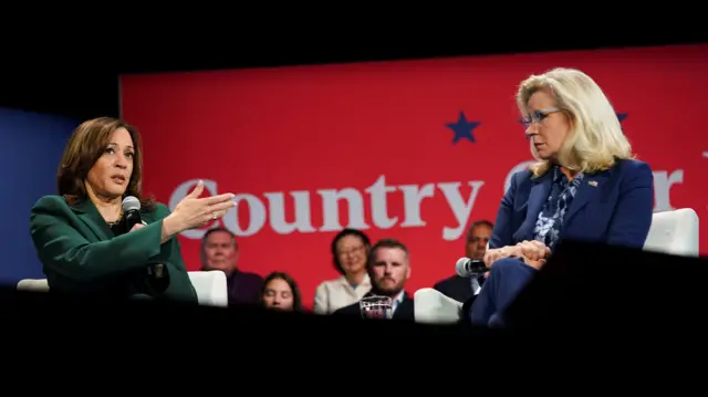 Harris in dark green suit speaks as she shares the stage Cheney in blue suit and patterned shirt, both are seated. Audience members sitting behind them, blurred, in front of red background