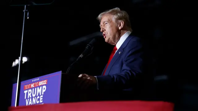 Trump stands at a podium where it reads: Trump Vance. He is wearing a blue suit with a red tie