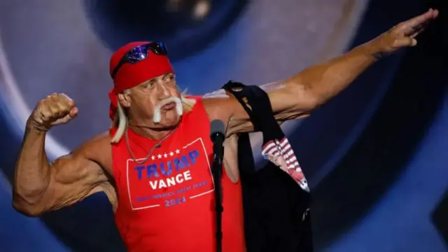 Hulk Hogan wearing a 'Trump-Vance' red tank top at this summer's Republican National Convention. Remnants of a ripped up black t-shirt hang loose off his left arm, which is extended sideways. His right arm is bent and hand is in a fist position.