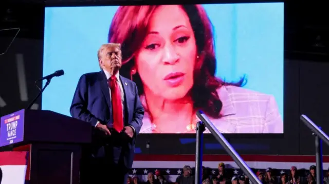 Donald Trump on stage in front of a screen showing Kamala Harris