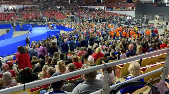 A wideshot of the stadium in Wisconsin is seen, with hundreds of people standing in their seats. The stage is empty but is adorned in bright blue carpet