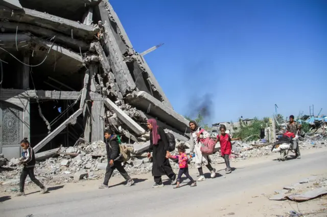 Children and a woman walking with backpacks in front of rubble. Behind them, a person pushing a filled cart