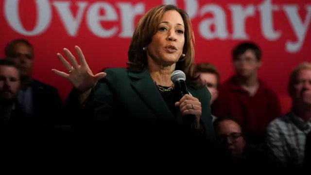 Kamala Harris is wearing a green blazer and gesturing in front of a blurred red background. Five men and a woman are sitting behind her but faces are blurry