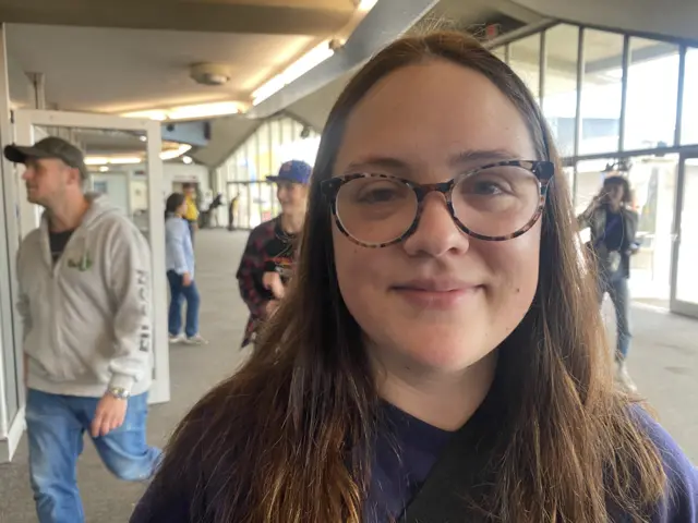 A young woman, Emily Kingman, stands in the middle of a bright convention centre. She has brown hair and glasses on.