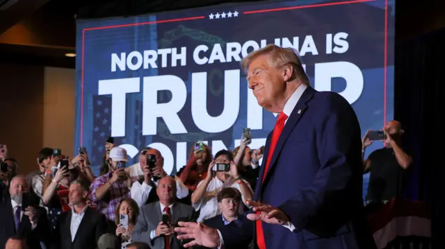 Trump in dark blue suit, white shirt and red tied, he has American flag pin on left lapel. Behind him is a group of evengelical voters taking pictures. In background is large screen reading North Carolina is Trump country