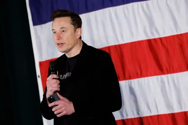 Elon Muks in black jacket and black t-shirt. He's holding mic in right hand and a phone in his left hand as he speaks. Large American flag in background