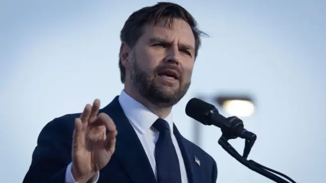 JD Vance, in a blue suit and tie, gestures with his right hand as he speaks at a campaign rally in Michigan