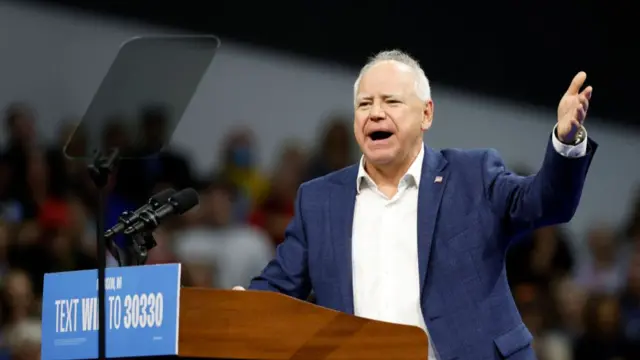 Tim Walz stands at a podium and gestures with his left hand. He is dressed in a blue suit jacket and white dress shirt