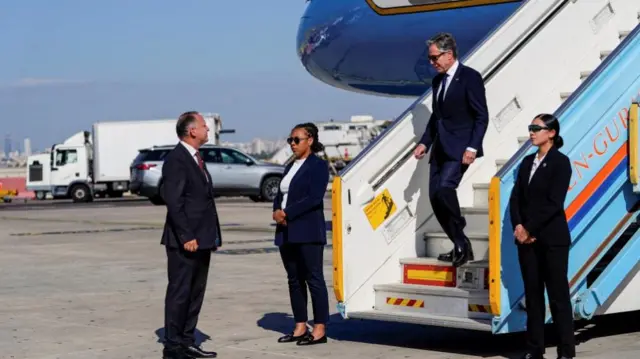 Antony Blinken walks down stairs of plane onto to runway. Two security guards stand at either side of the bottom of the stairs, and a man is standing ready to great him. All are dressed in dark suits