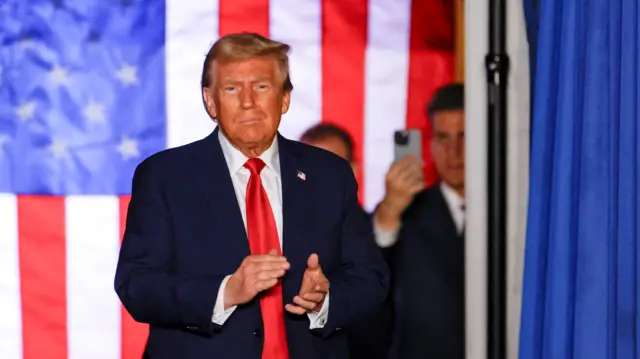 Trump in dark blue, white shirt and bright red tie, claps in front of a US flag. A man to his left holds a phone up in the blurred background