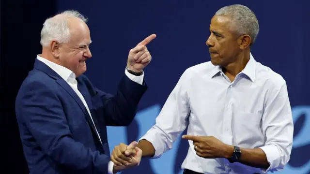 Tim Walz and Barack Obama hold hands and Walz points at Obama as they meet on stage