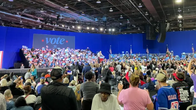 A crowd of people in a large stadium with a sign saying 'vote' in the background