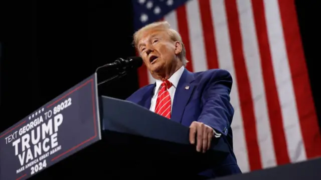 Trump speaking at a podium in Atlanta, Georgia during a rally. He's in blue suit, red tie with white stipes and American flag pin on left lapel, American flag hanging behind him