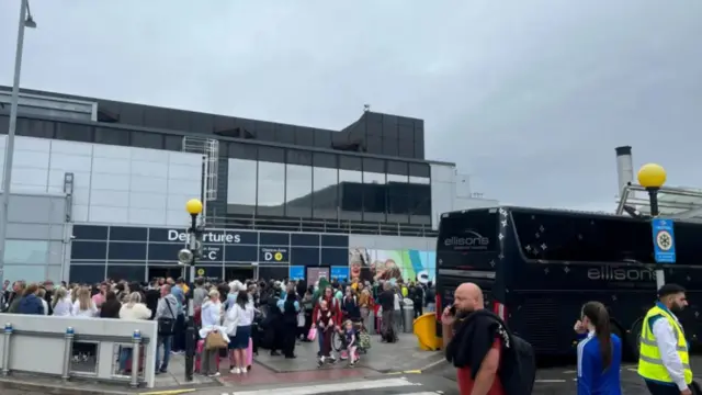 People standing outside an airport with some wearing yellow jackets