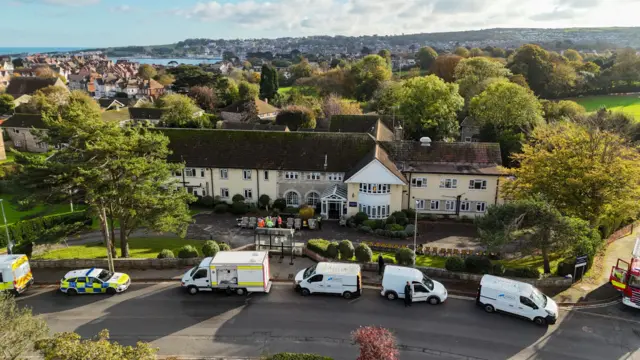 Drone image of the two-storey, cream-coloured care home with a line of police, fire and gas vehicles parked outside. There are trees behind the building and the town of Swanage in the distance beyond.