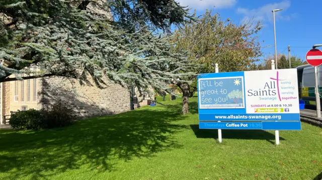 Outside All Saints Church, a sign for the church on a patch of green grass