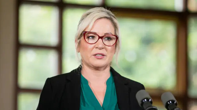 Michelle O'Neill stands in front of two black microphones. She is wearing her blonde hair tied back, with two strands framing her face. She is wearing red-rimmed glasses, a black blazer and a green shirt.