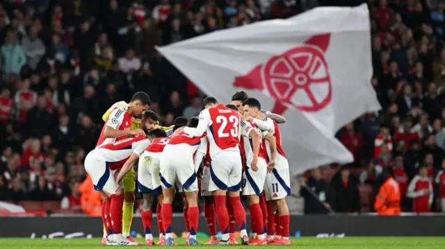 Arsenal players in a huddle with a huge Gunners flag in the background