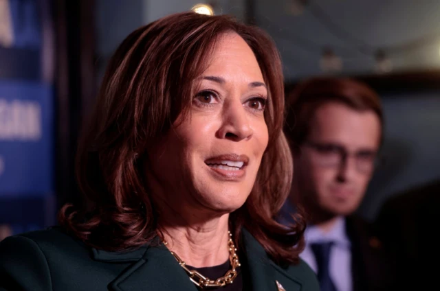 Kamala harris close us. She's in a green suit with black blouse and a chunky gold chain necklace