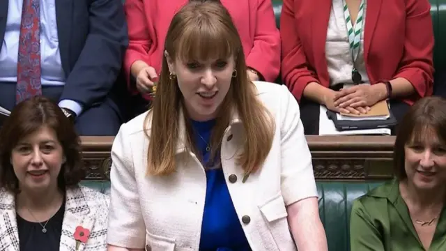 Deputy PM Angela Rayner at PMQs,. She's wearing white short-sleeved jacket with two side pockets and brown buttons, blue shirt underneath. Rachel Reeves sitting at her left, Lucy Powell to her right