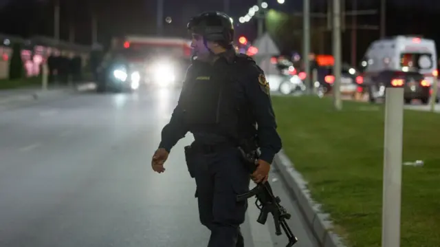 An armed policeman walks near to the scene, with emergency vehicles in the background