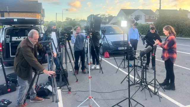 Members of the press stand in a car park with multiple lights, cameras and two vans in the background