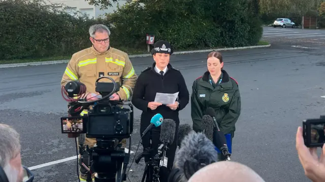 Darren Langdown from the fire service stands next to ch supt Heather Dixey and Susan Thomas from the ambulance service, they are standing in front of cameras and lights in a car park for a press conference