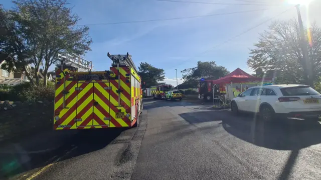 A residential road with a fire engine parked on the left, a white car parked on the right next to a red marquee and ambulance cars in the background