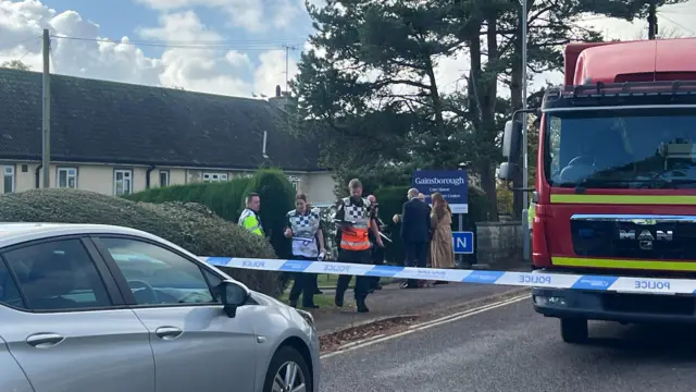 A blue and white police tape goes across a section of road, in front of a fire engine. A sign for Gainsborough Care Home can be seen behind the fire engine, it's navy blue and white. There are a group of three mean and a woman crowded in a circle in front of the sign, a bit further in front of them a man and woman look like they're leaving the care home. They are wearing black and white checked high vis vest and a police officer with a yellow high vis coat on is behind them