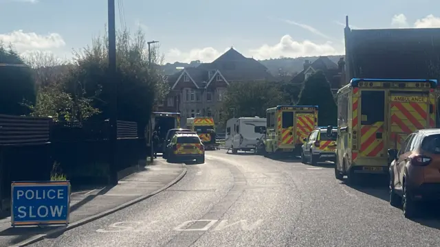 A police slow sign sits on the road to the left of the image as ambulances and police vehicles line either side of a residential street leading up to the care home