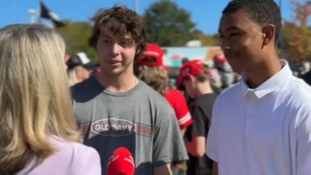 Two men speak with a reporter holding a microphone at an outdoors event