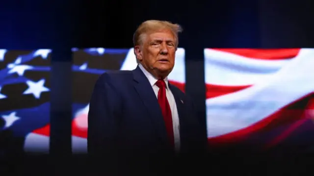 Donald Trump in black suit and red tie stands in front of maxi screens showcasing sections of the US flag