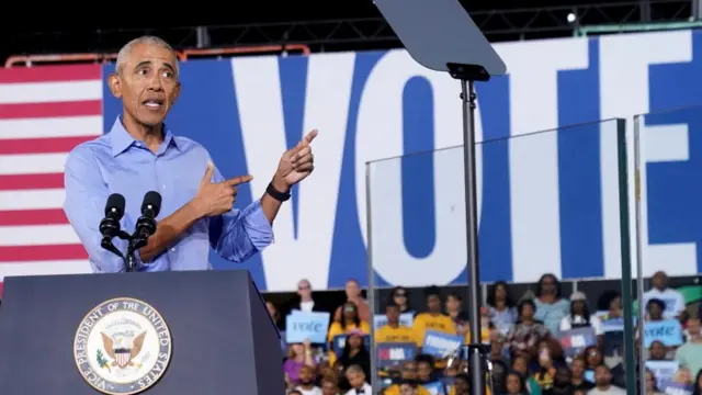 Barack Obama speaks on stage in a blue shirt with the words “Vote” visible in the background.