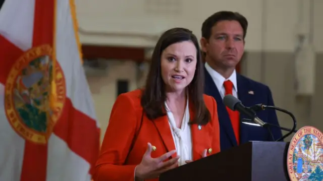 Ashley Moody speaks at a podium, with Florida governor Ron DeSantis at her right and the Florida flag at her left.